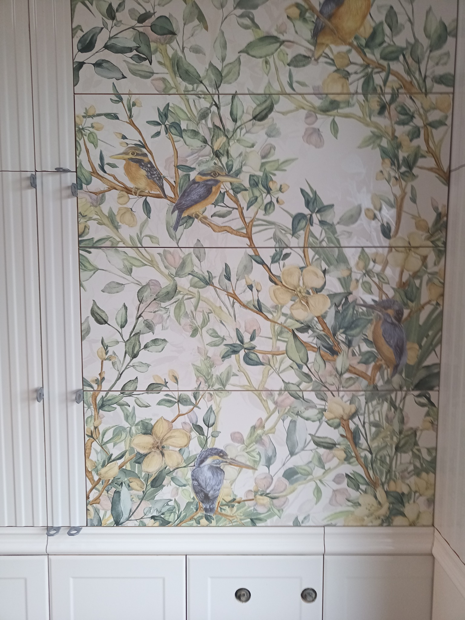 Ściana wyłożona płytkami ceramicznymi z motywem akwarelowych ptaków i kwitnących gałęzi, uzupełniona białymi szafkami z frezowanymi frontami.