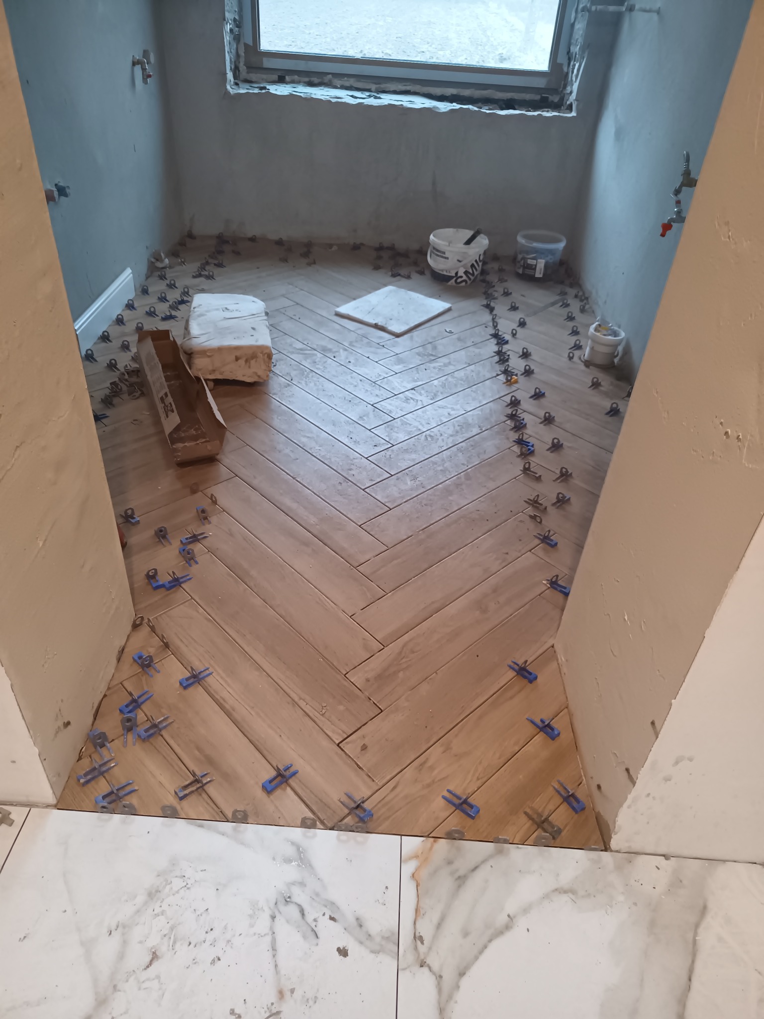 Układanie drewnopodobnych płytek w jodełkę w łazience w trakcie prac wykończeniowych, widoczne niezagruntowane ściany, paczka kleju, wiaderka i niebieskie klipsy poziomujące.