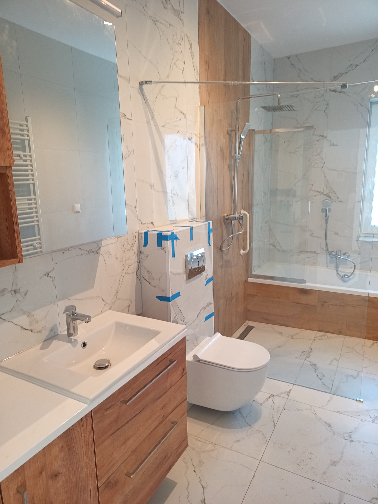 Nowoczesna łazienka z kabiną prysznicową, umywalką z drewnianą szafką i toaletą. Marmurowe płytki na ścianach i podłodze. Część elementów zabezpieczona taśmą.