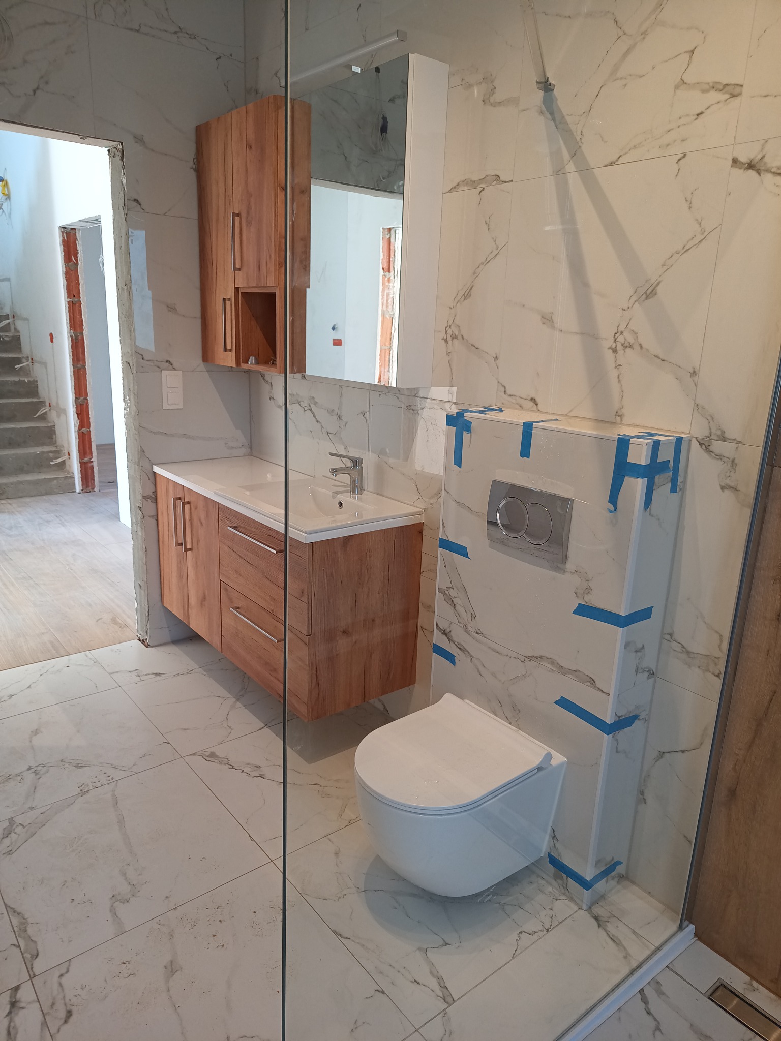 Nowoczesna łazienka z drewnianą szafką pod umywalką, marmurową płytką i podwieszaną toaletą z niebieską taśmą zabezpieczającą. Widok na niedokończone schody.