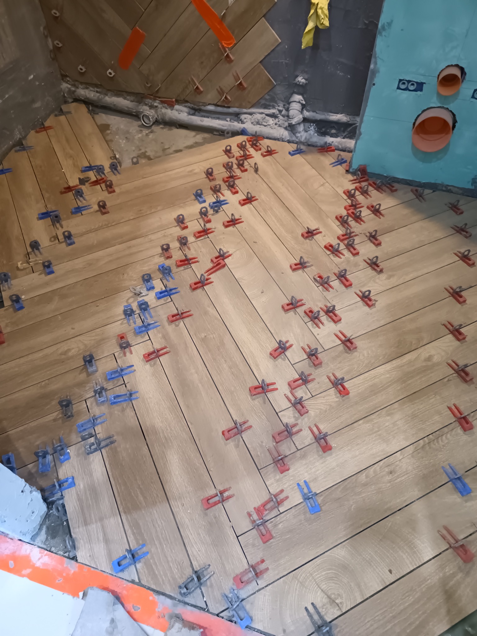 Układanie drewnopodobnych płytek na podłodze w łazience, z użyciem czerwonych i niebieskich klipsów poziomujących. Widoczne przygotowane ściany i instalacje.
