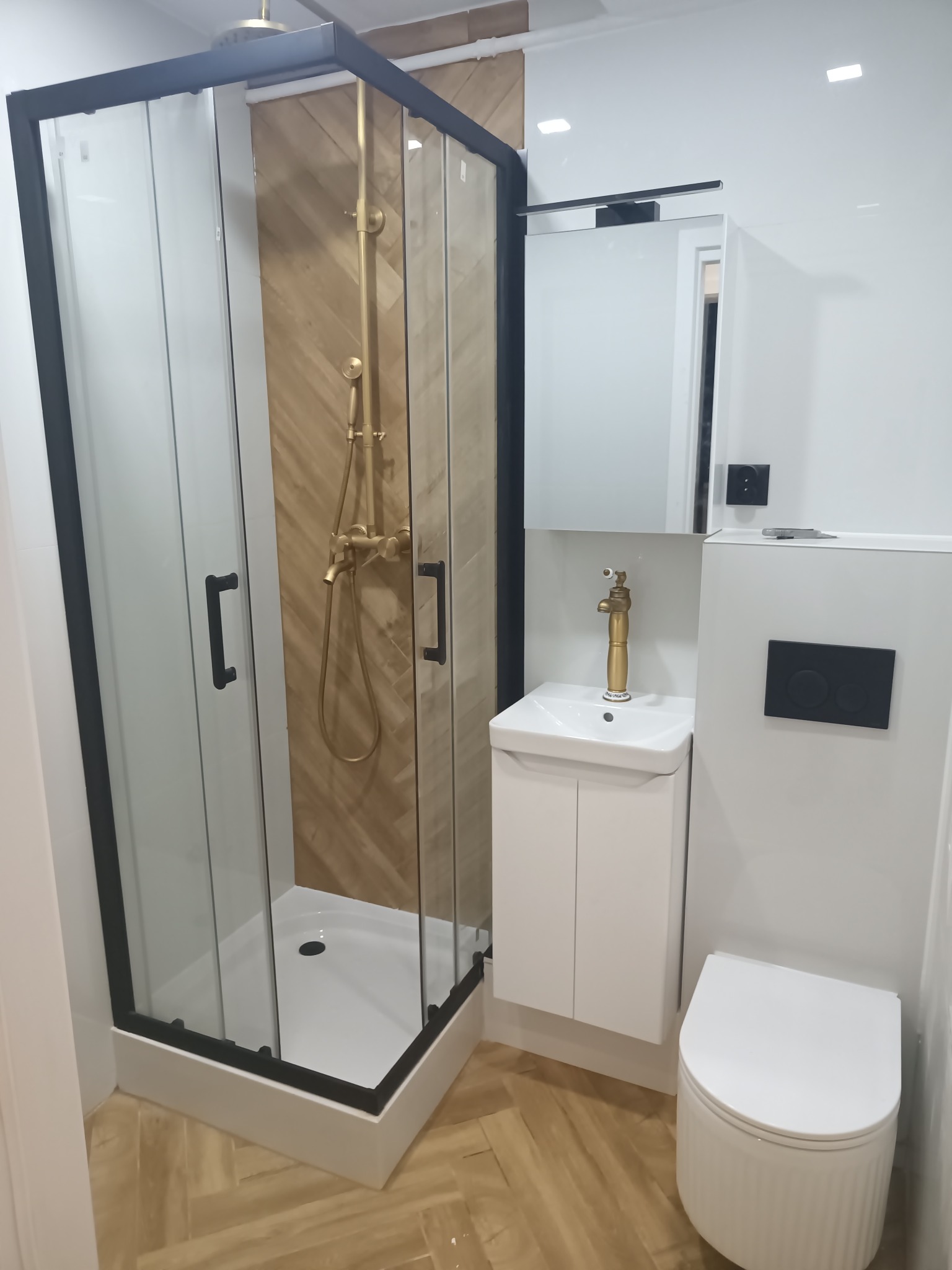 Nowoczesna łazienka z kabiną prysznicową w czarnej ramie, złotą armaturą i białymi meblami, wykończona w jasnych barwach z drewnopodobną podłogą.