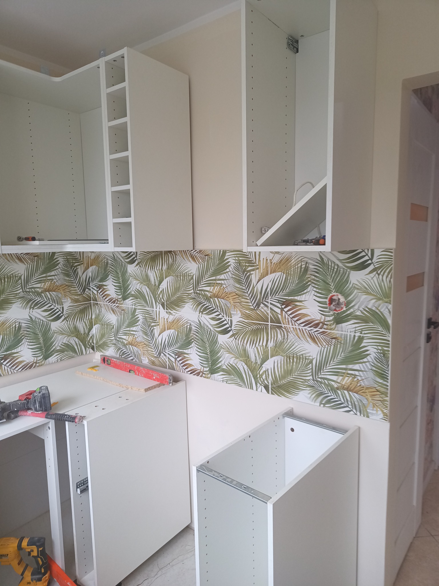 Montaż białych szafek kuchennych na tle ściany z motywem liści, widoczne narzędzia i poziomica, etap prac wykończeniowych.