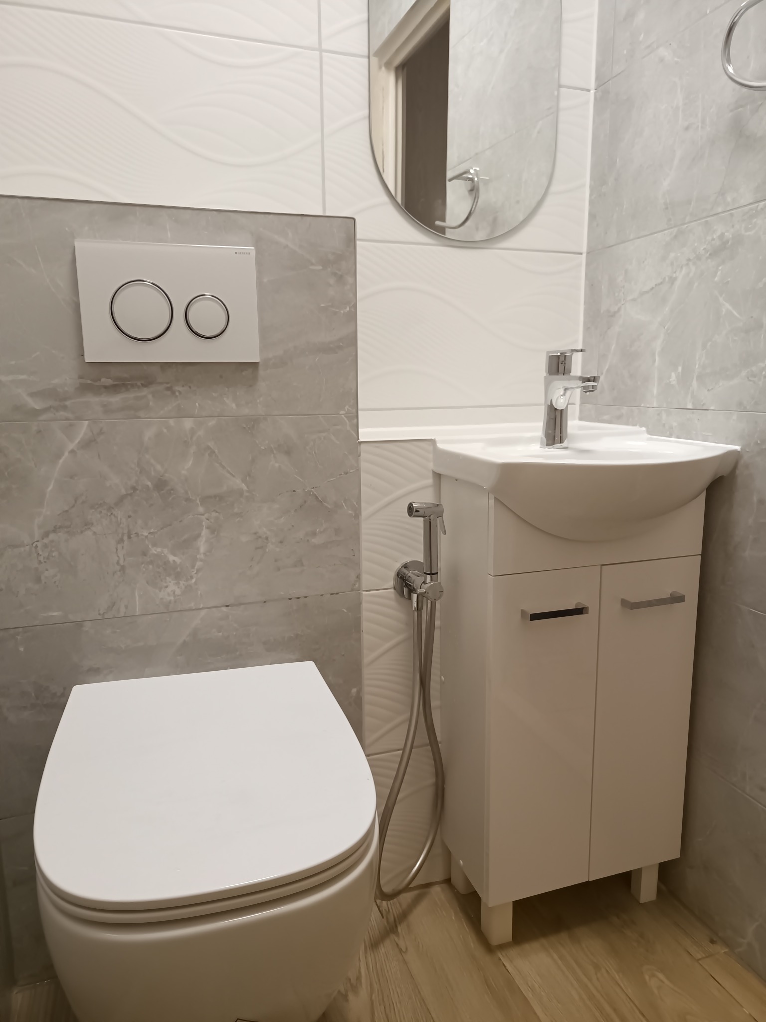 Nowoczesna łazienka po remoncie: biała umywalka z szafką, toaleta z przyciskiem Geberit, bidetka i lustro na tle szarych płytek imitujących marmur.