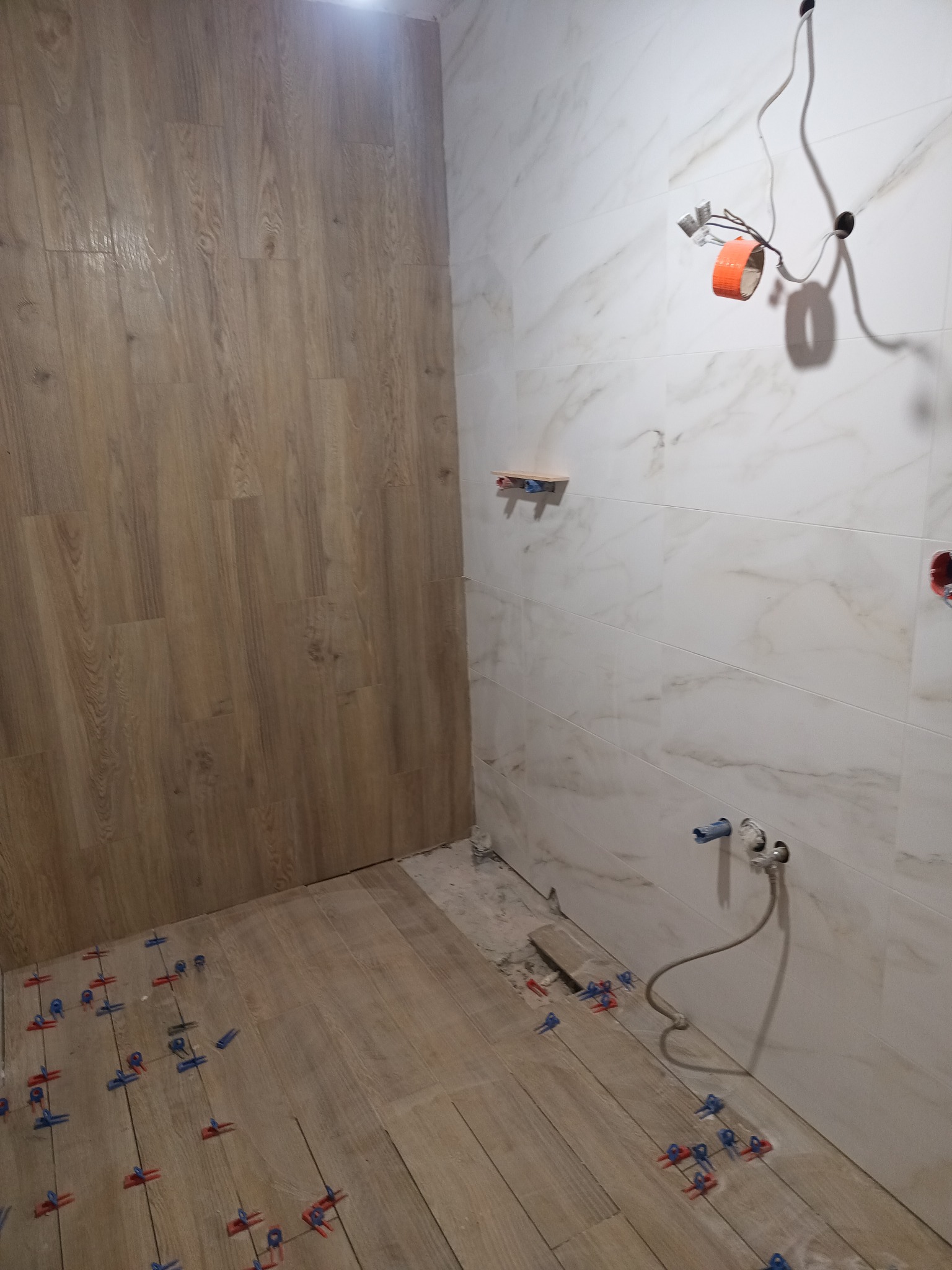 Układanie płytek imitujących drewno na podłodze i marmurowych na ścianach w trakcie prac. Widoczne krzyżyki dystansowe i wystające kable.