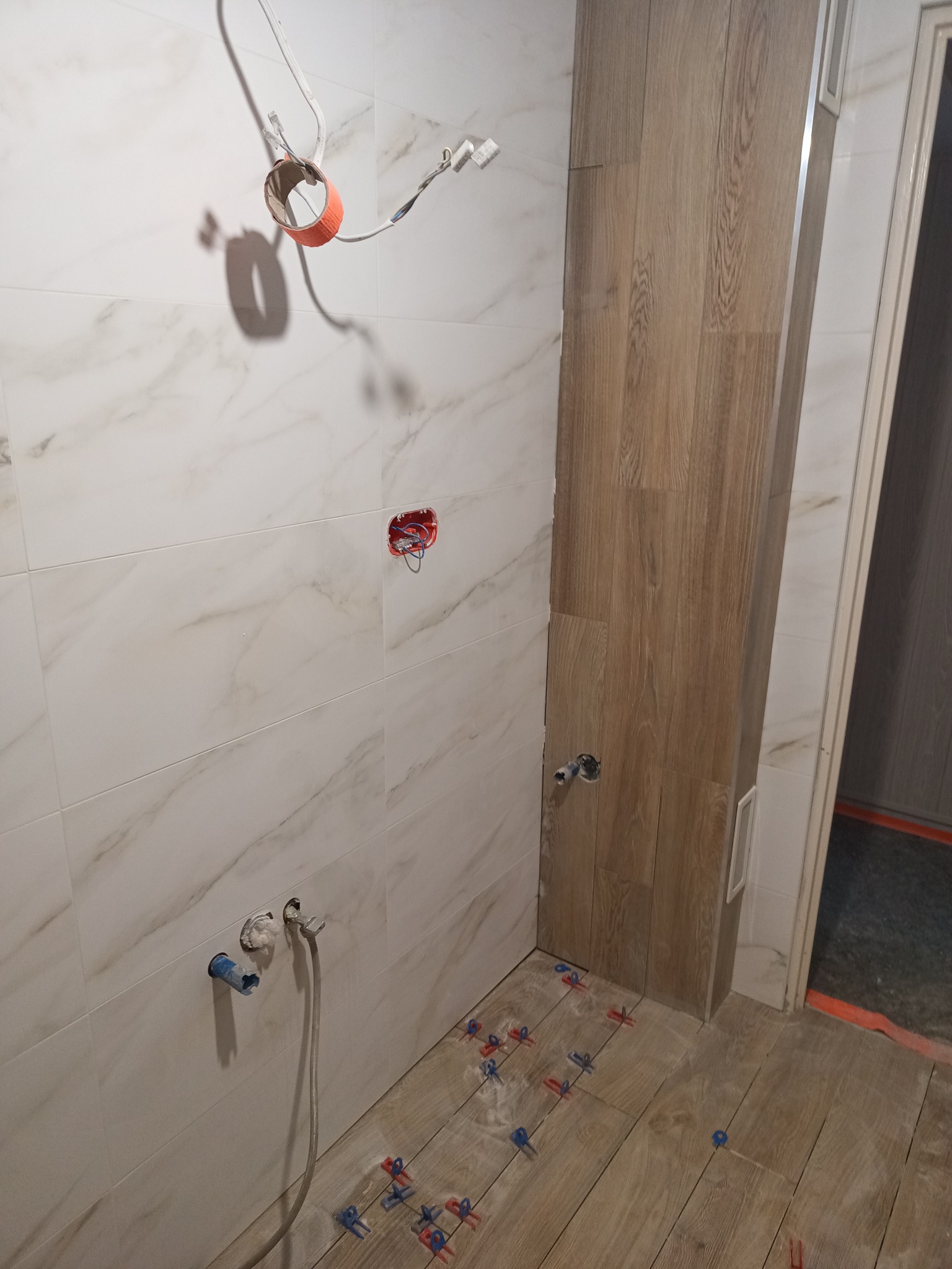 Łazienka w trakcie prac: białe płytki imitujące marmur na ścianach, drewnopodobne płytki na podłodze, widoczne instalacje i akcesoria montażowe.