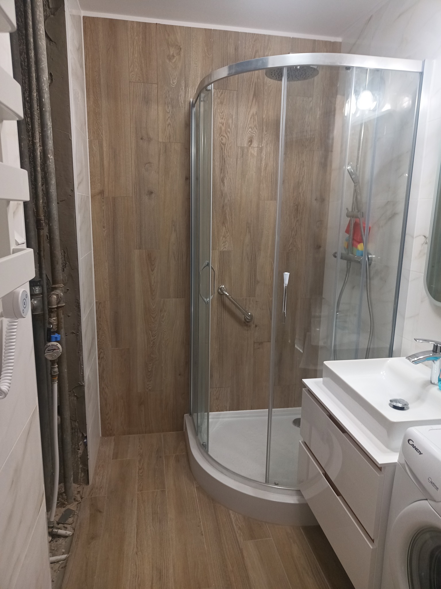 Wykończona łazienka z kabiną prysznicową, drewnopodobnymi płytkami na ścianach i podłodze. Widoczne rury grzewcze przed zabudową. Umywalka z szafką i pralka.