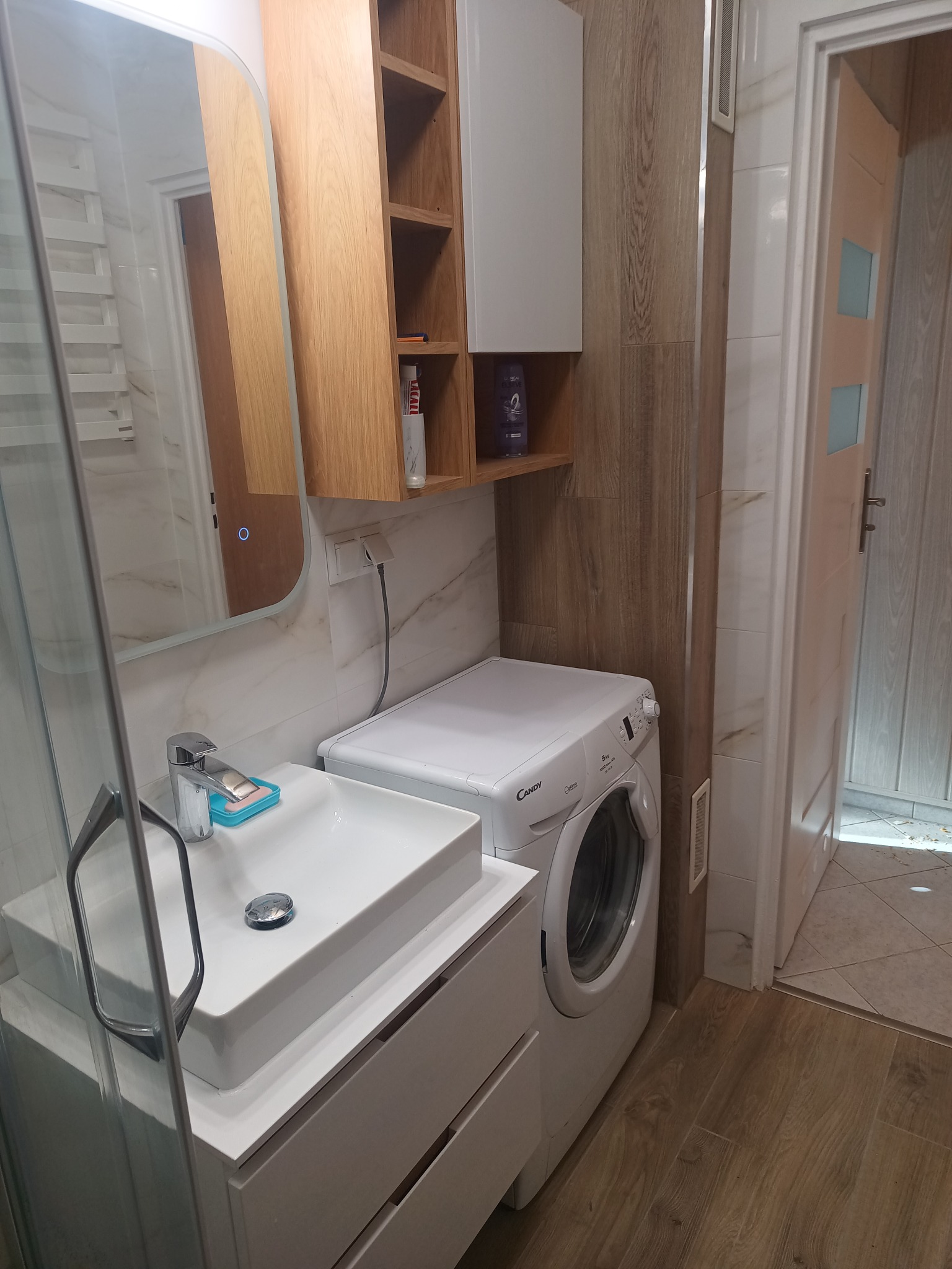 Nowoczesna łazienka z umywalką nablatową, pralką Candy i drewnianymi szafkami. Lustro z podświetleniem LED. Płytki imitujące drewno na ścianie.