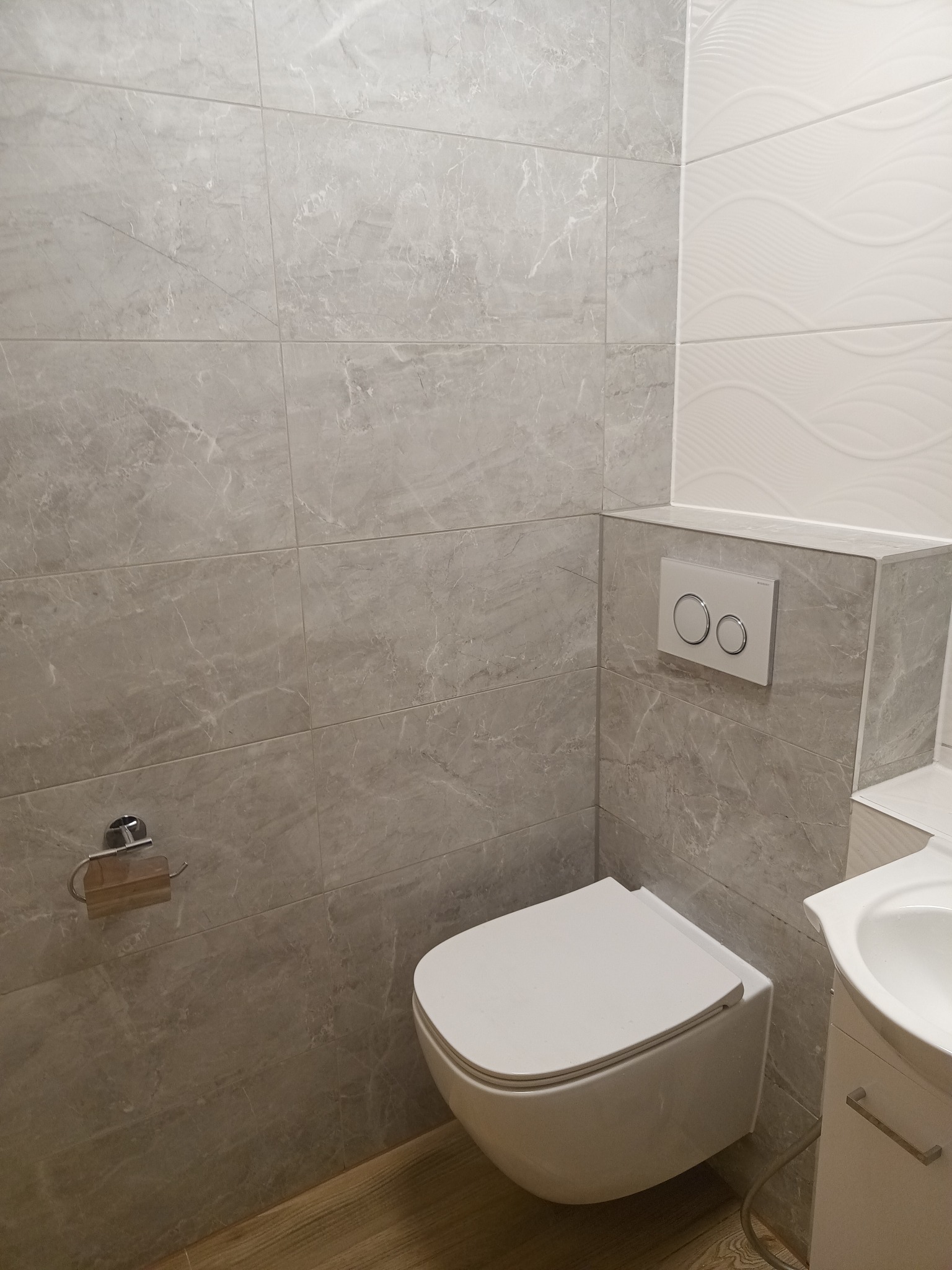 Nowoczesna łazienka z szarymi płytkami imitującymi marmur, podwieszana toaleta i umywalka. Na ścianie widoczny przycisk spłukujący. Minimalistyczny design.