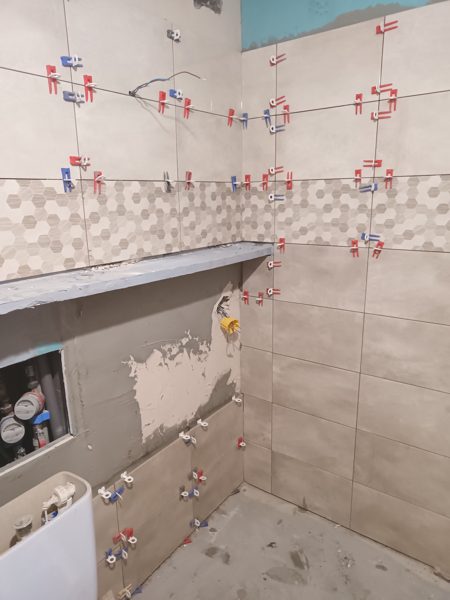 Układanie beżowych płytek z dekoracyjnym pasem w kształcie plastra miodu w łazience. Widoczne klipsy poziomujące i fragment instalacji wodnej.