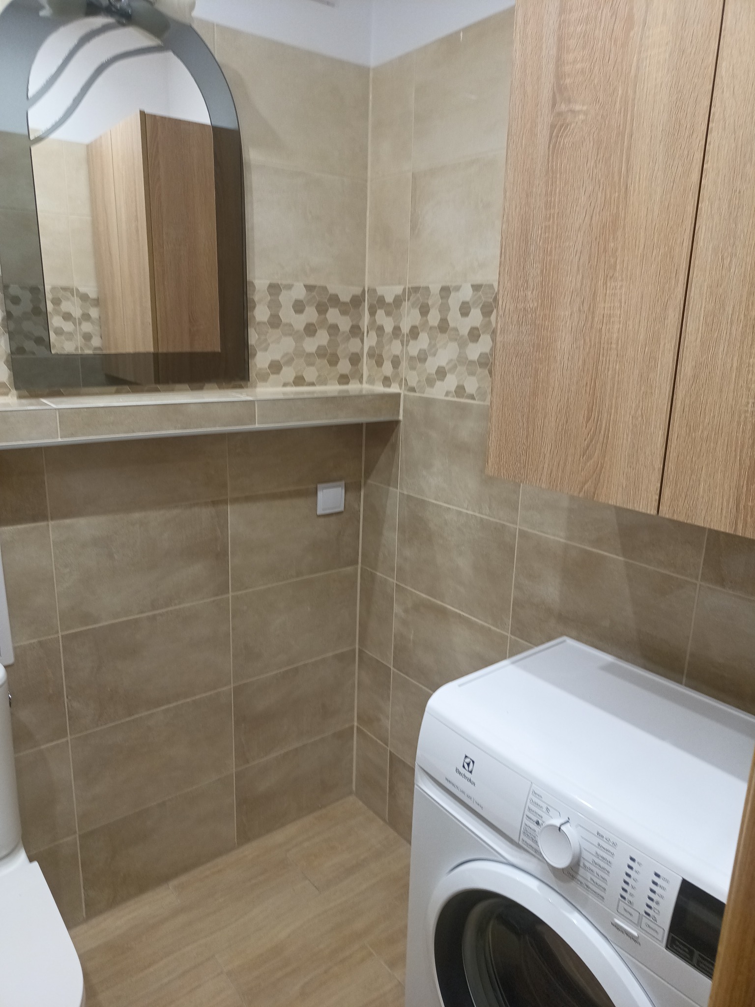 Jasna łazienka po remoncie: beżowe płytki, szafka z drewnianym frontem, pralka Electrolux i lustro w kształcie łuku. Ukończone wykończenie wnętrza.