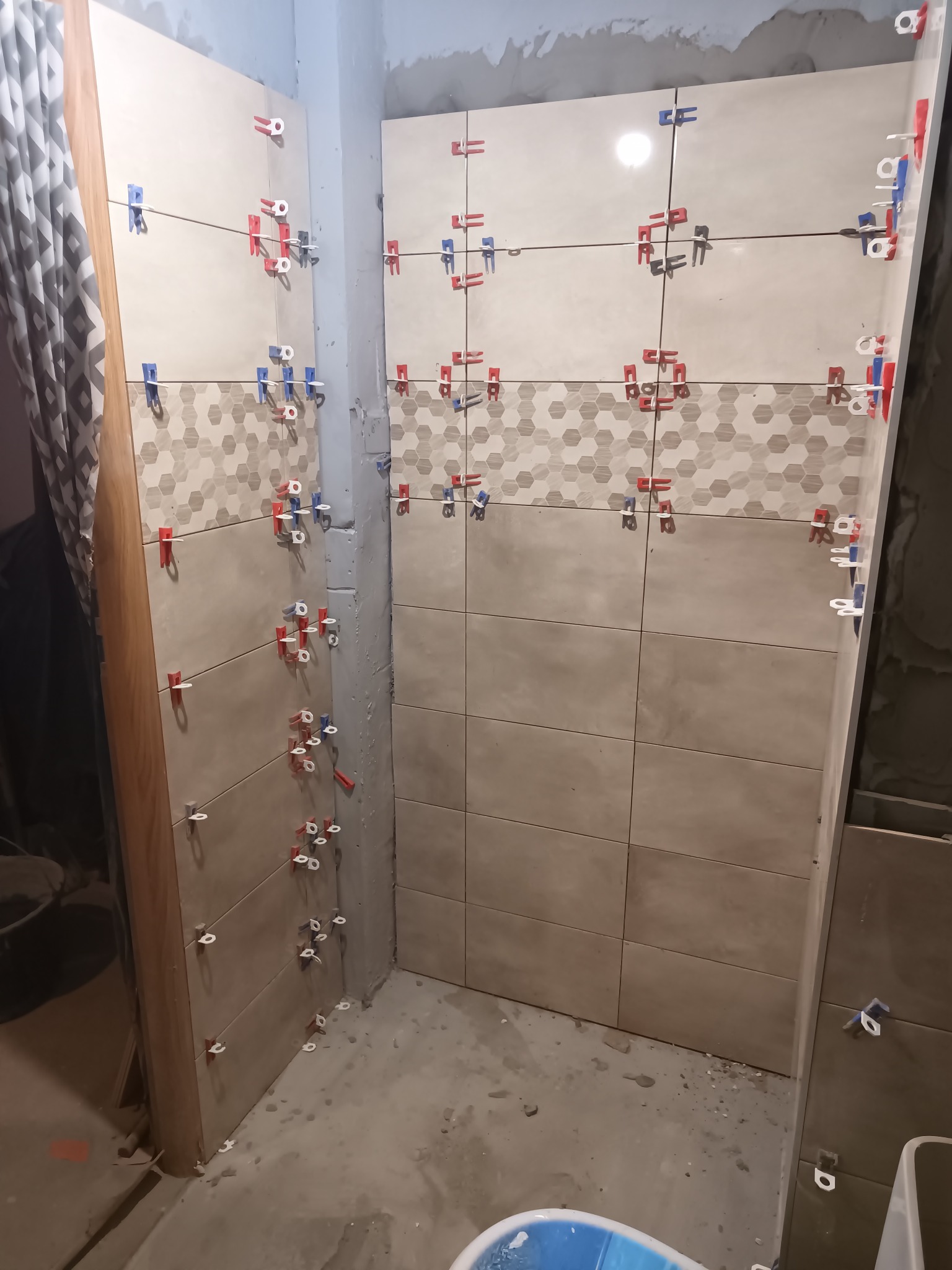 Układanie płytek w łazience z użyciem klipsów poziomujących. Widoczne płytki w odcieniach beżu z dekoracyjnym pasem w geometryczny wzór. Prace w toku.