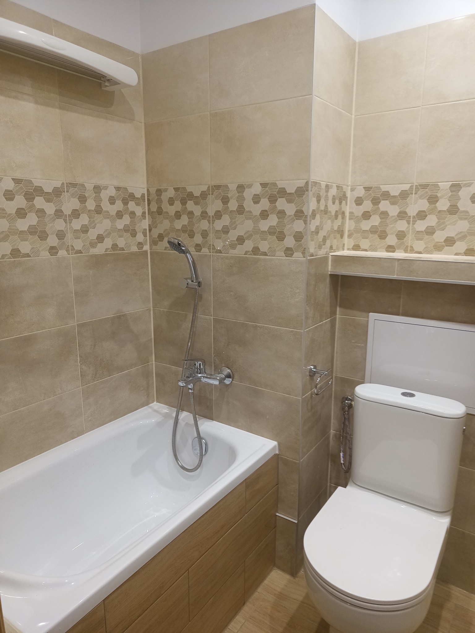 Jasna łazienka z beżowymi płytkami, dekoracyjnym pasem z motywem plastra miodu, białą wanną i toaletą. Minimalistyczny design, ciepłe oświetlenie.