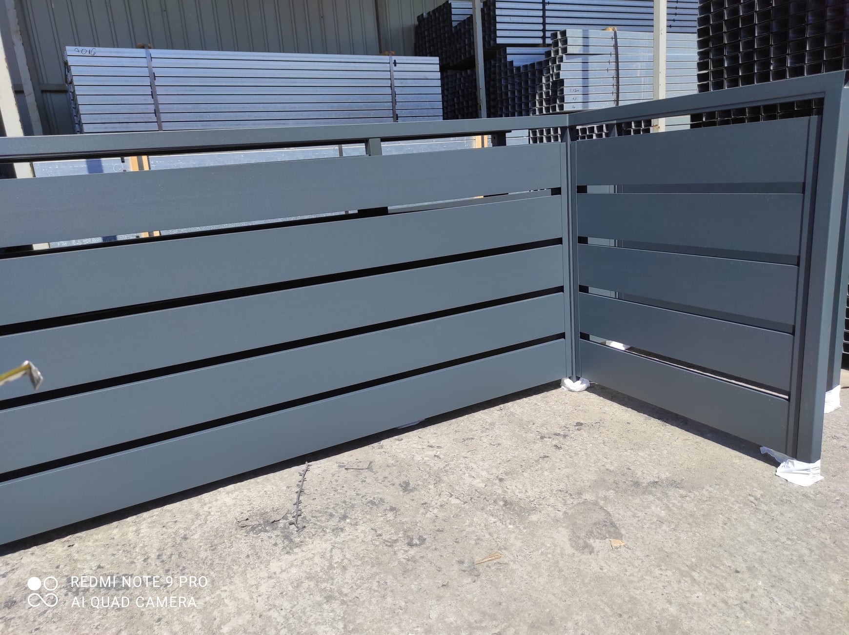 Nowoczesna, antracytowa brama wjazdowa z poziomymi panelami, widoczna w trakcie montażu, z materiałami składowanymi w tle.