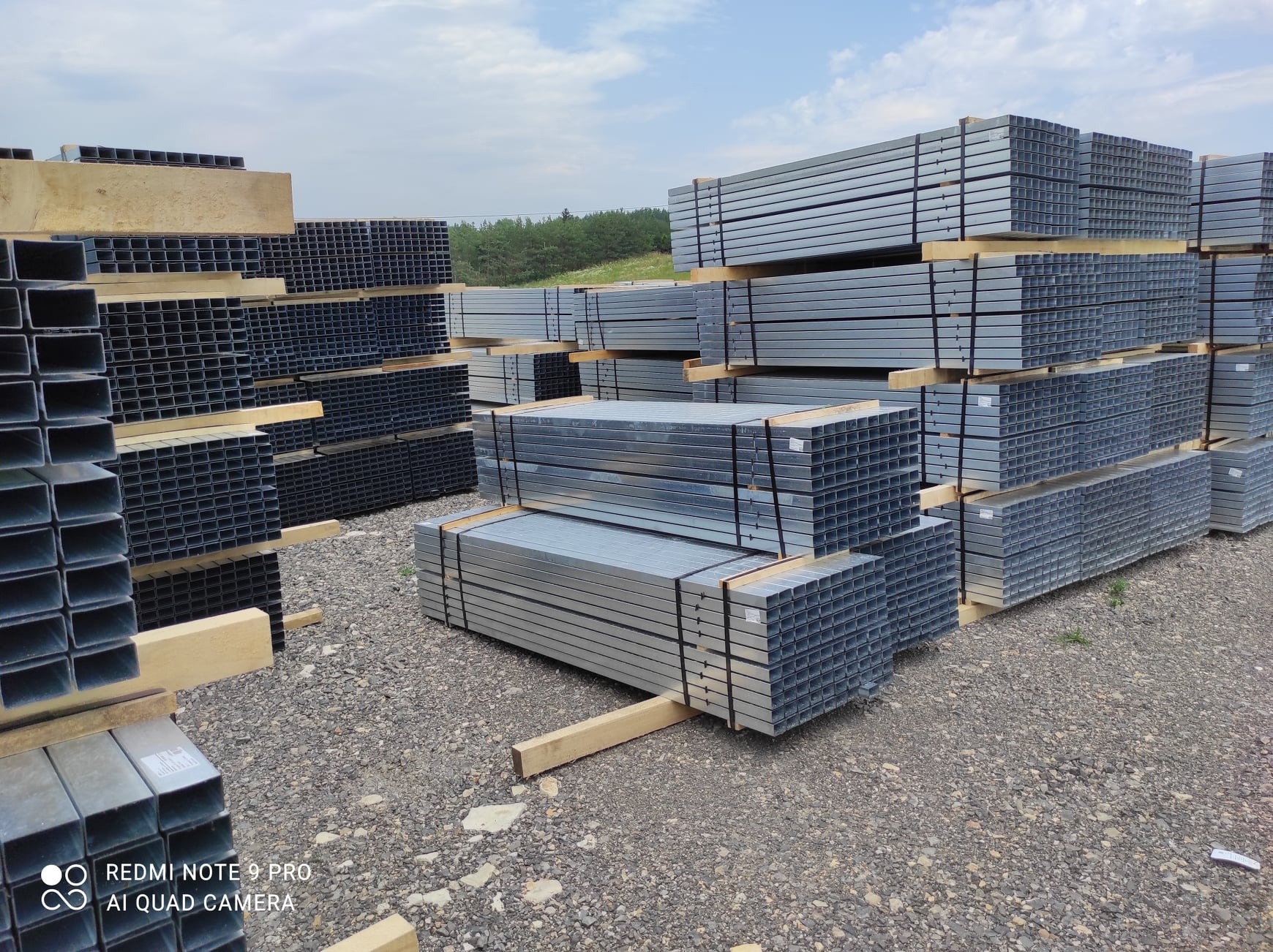 Stosy ocynkowanych profili stalowych C i kwadratowych, ułożone na drewnianych belkach na żwirowym podłożu, widok z zewnątrz.