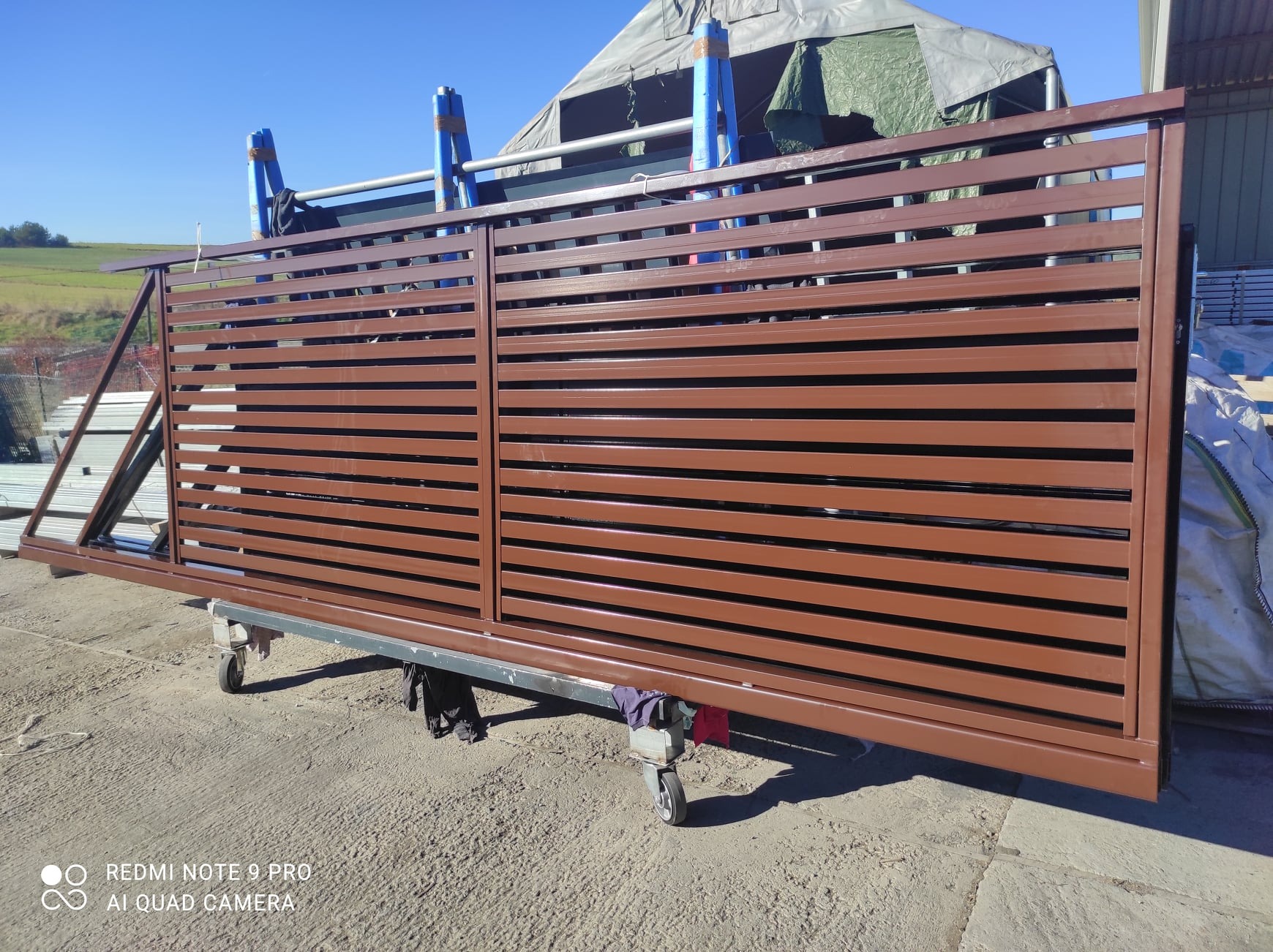 Brązowa, przesuwna brama wjazdowa z poziomymi panelami, na wózku transportowym w Trzyciążu, gotowa do montażu.
