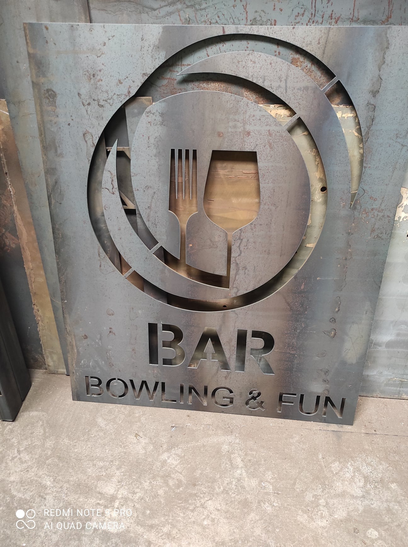 Metalowy szyld z wyciętym motywem kieliszka, widelca i napisem 'BAR BOWLING & FUN', surowy wygląd, widoczne ślady obróbki.
