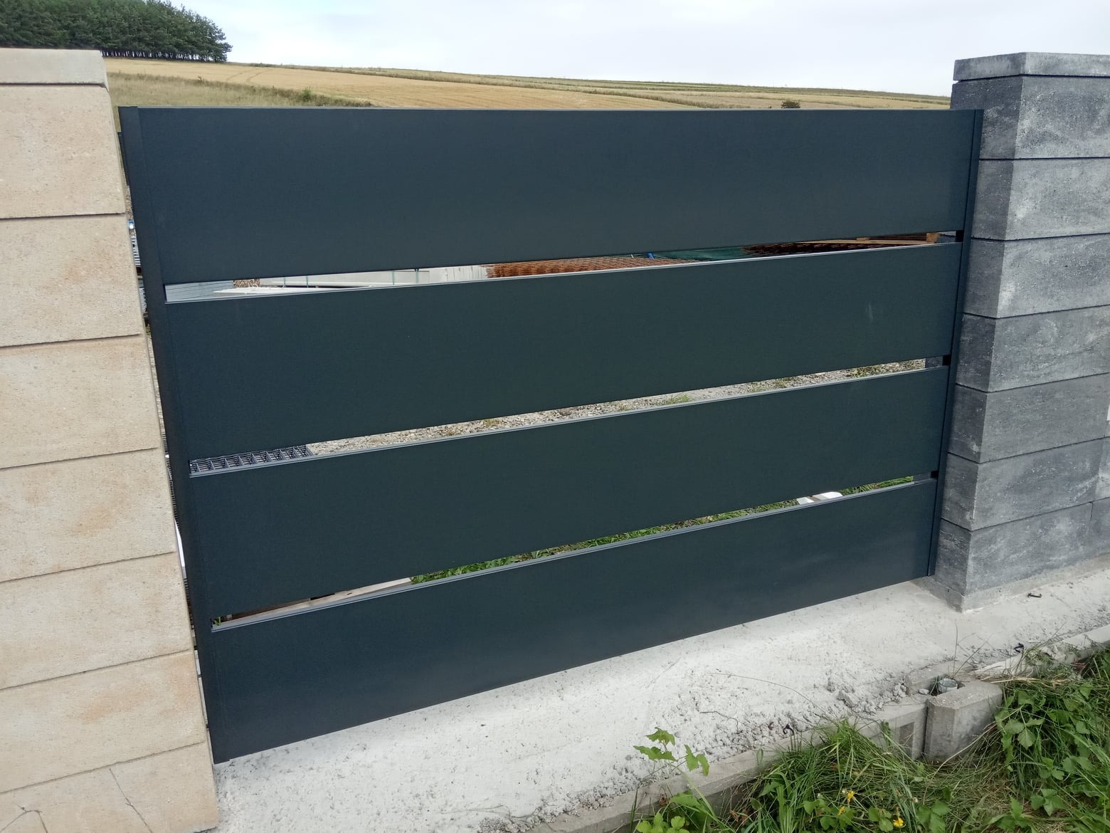 Nowoczesne ogrodzenie z poziomych, grafitowych paneli, osadzone między słupkami z szarej kostki betonowej. W tle widoczne pole i drzewa.