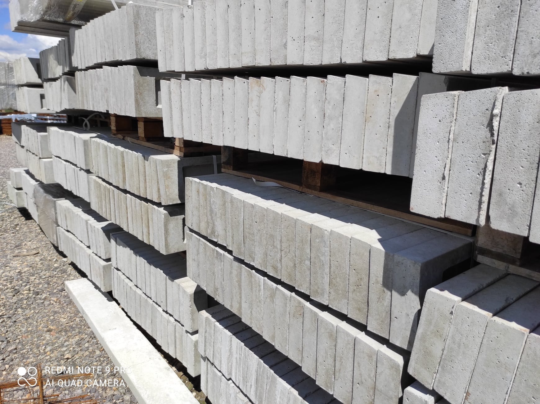 Stos szarych, betonowych słupków ogrodzeniowych ułożonych na paletach, widoczne drobne nierówności powierzchni betonu i szary żwir w tle.