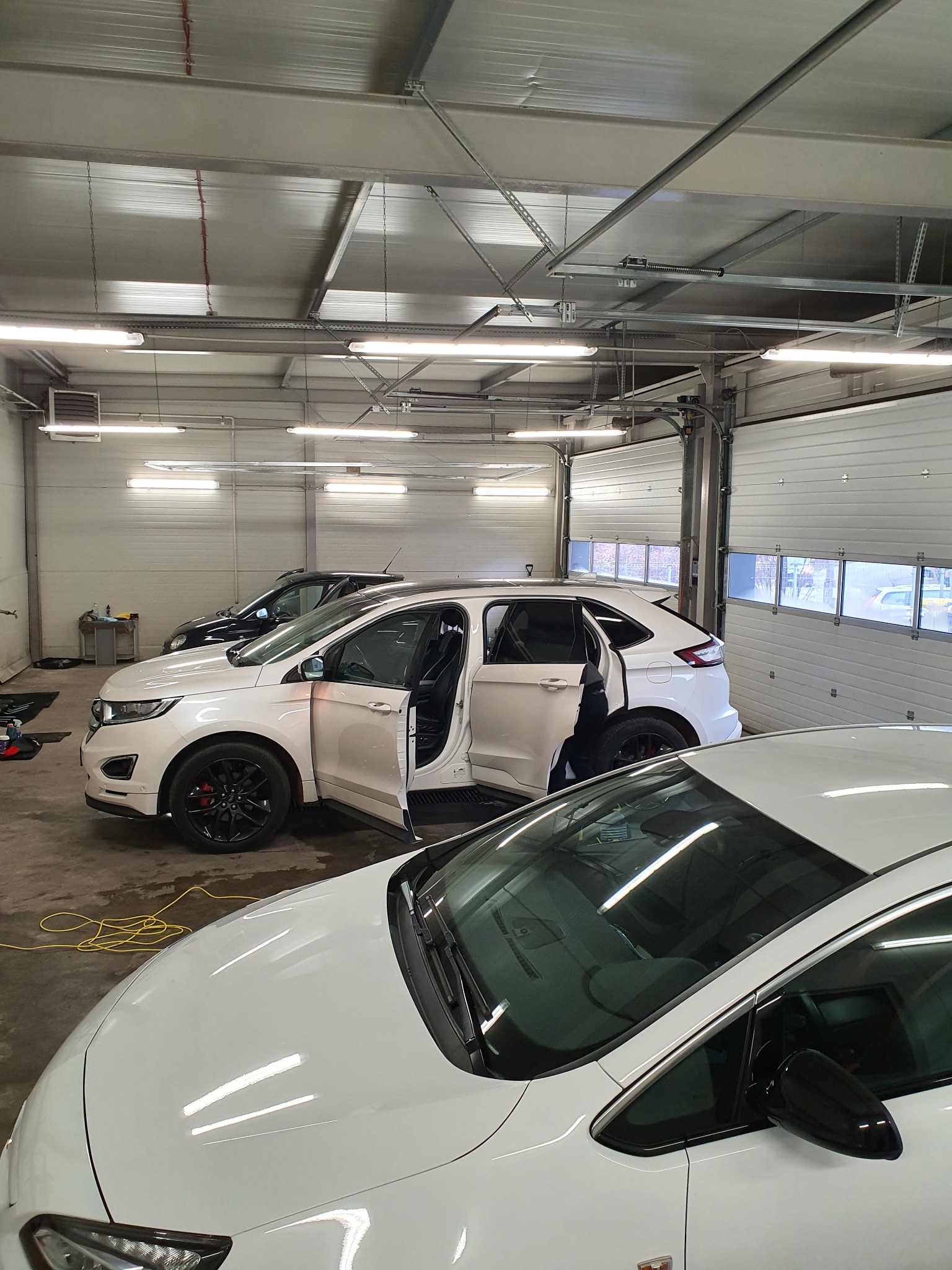 Wnętrze myjni samochodowej z trzema białymi autami, w tym Ford Edge z otwartymi drzwiami, widoczne okna w bramie garażowej i żółty kabel na podłodze.
