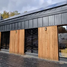 Nowoczesny dom z czarną blachą na dachu i elewacją z jasnego drewna, z zamontowanymi żaluzjami fasadowymi i lampami zewnętrznymi, widok z zewnątrz na tle jesiennego lasu.