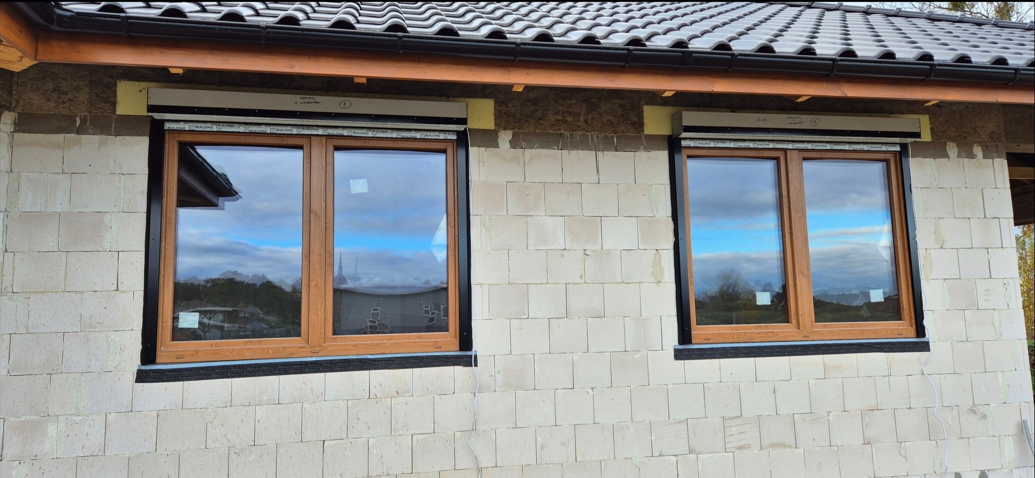 Dwa okna z drewnianą ramą i zamontowanymi systemami osłonowymi w kasecie, na surowej ścianie z białych bloczków, widoczne elementy montażu i izolacji.