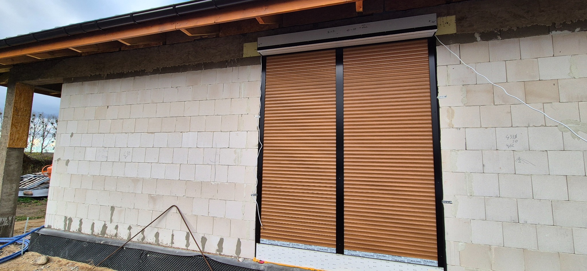 Zamknięte, brązowe rolety na oknie budynku w trakcie budowy, widoczne białe bloczki i czarne obramowanie okna.