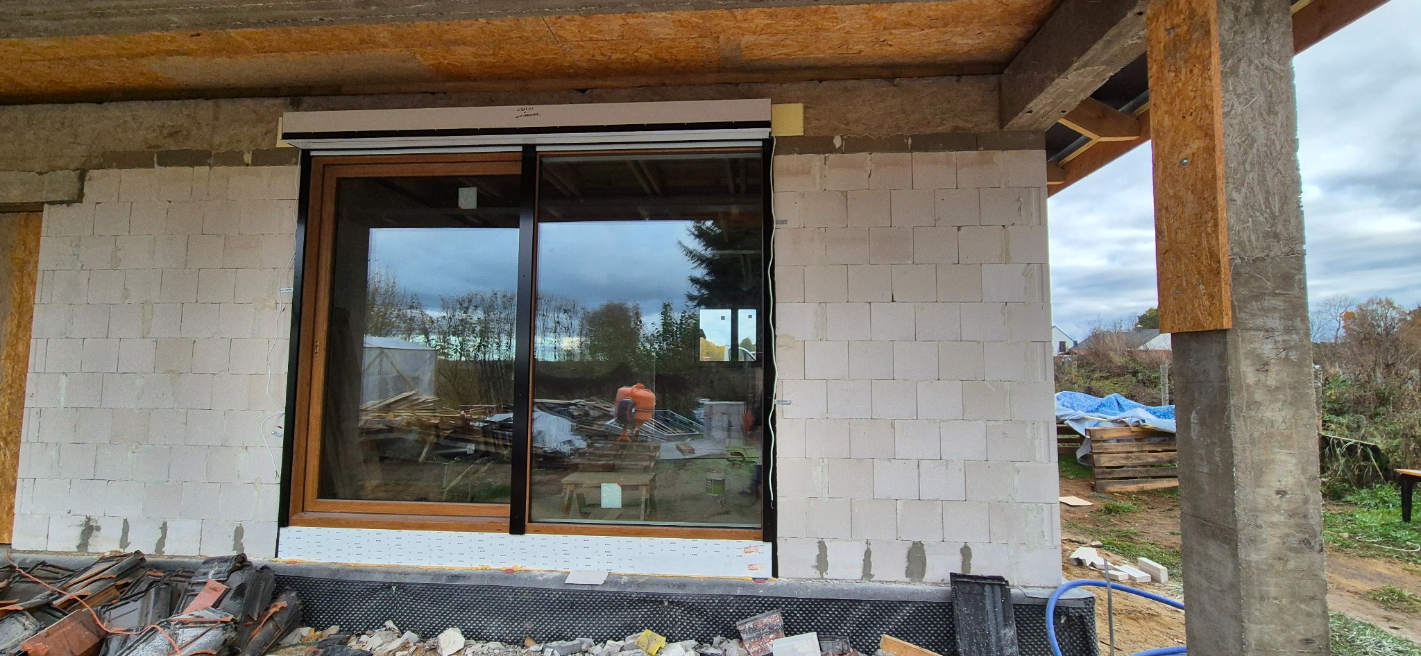 Montaż zewnętrznej rolety na oknie przesuwnym w surowym stanie budynku, widoczny mur z bloczków, elementy konstrukcyjne dachu oraz odbicie otoczenia w szybie.