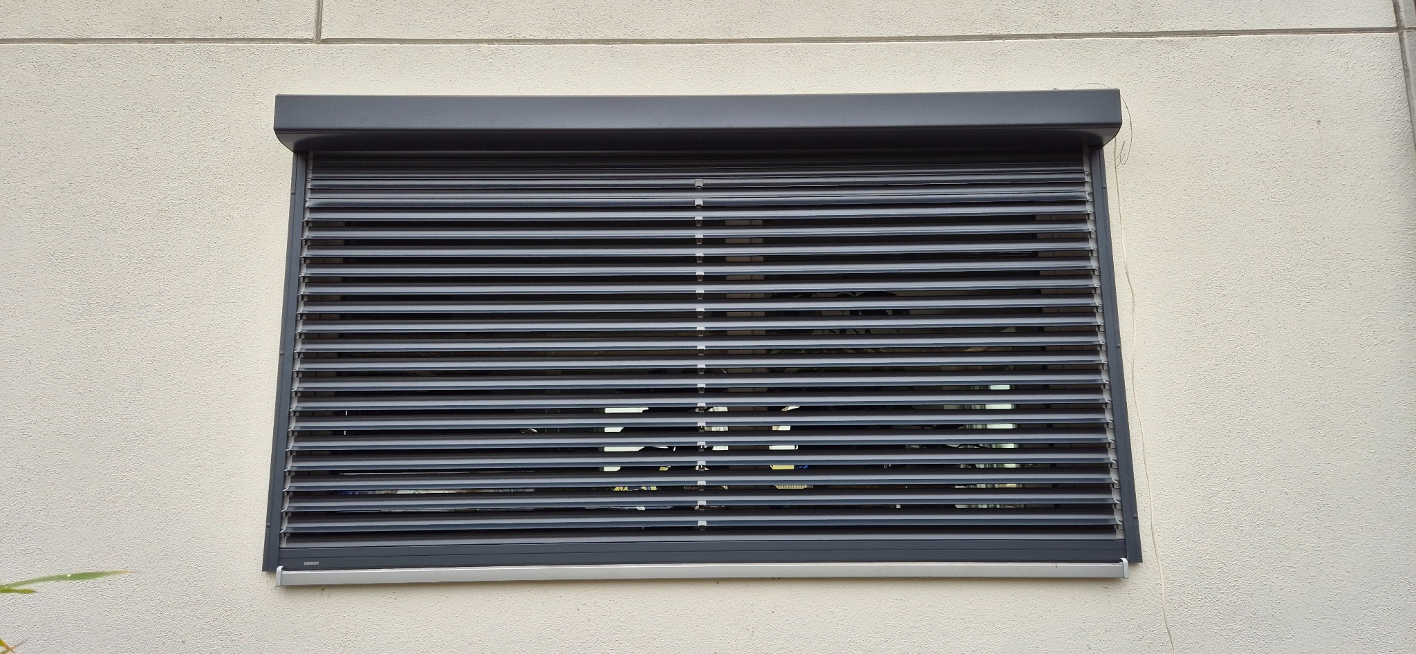 Szara żaluzja fasadowa zamontowana na elewacji budynku, widoczna z bliska, z delikatnym prześwitem światła przez lamele.