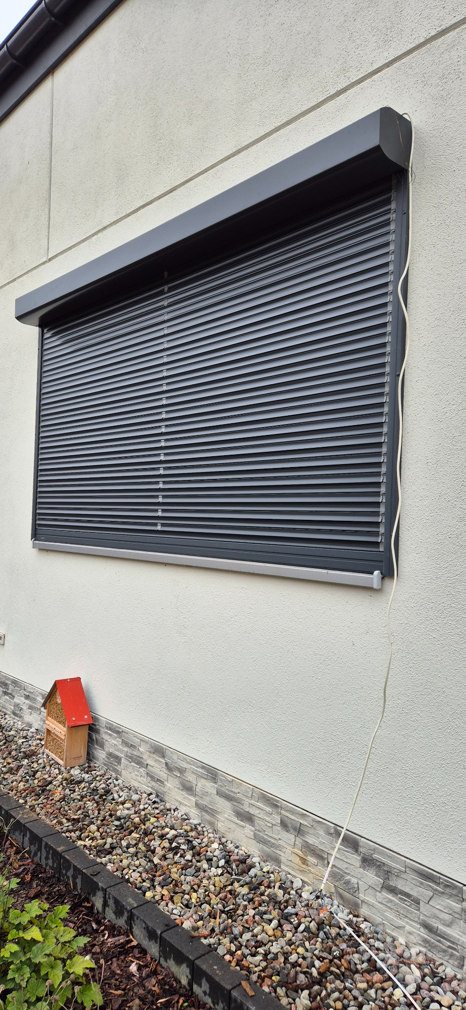 Zasłonięte okno szarą roletą zewnętrzną na elewacji domu, widoczna instalacja elektryczna i domek dla owadów w ogródku.