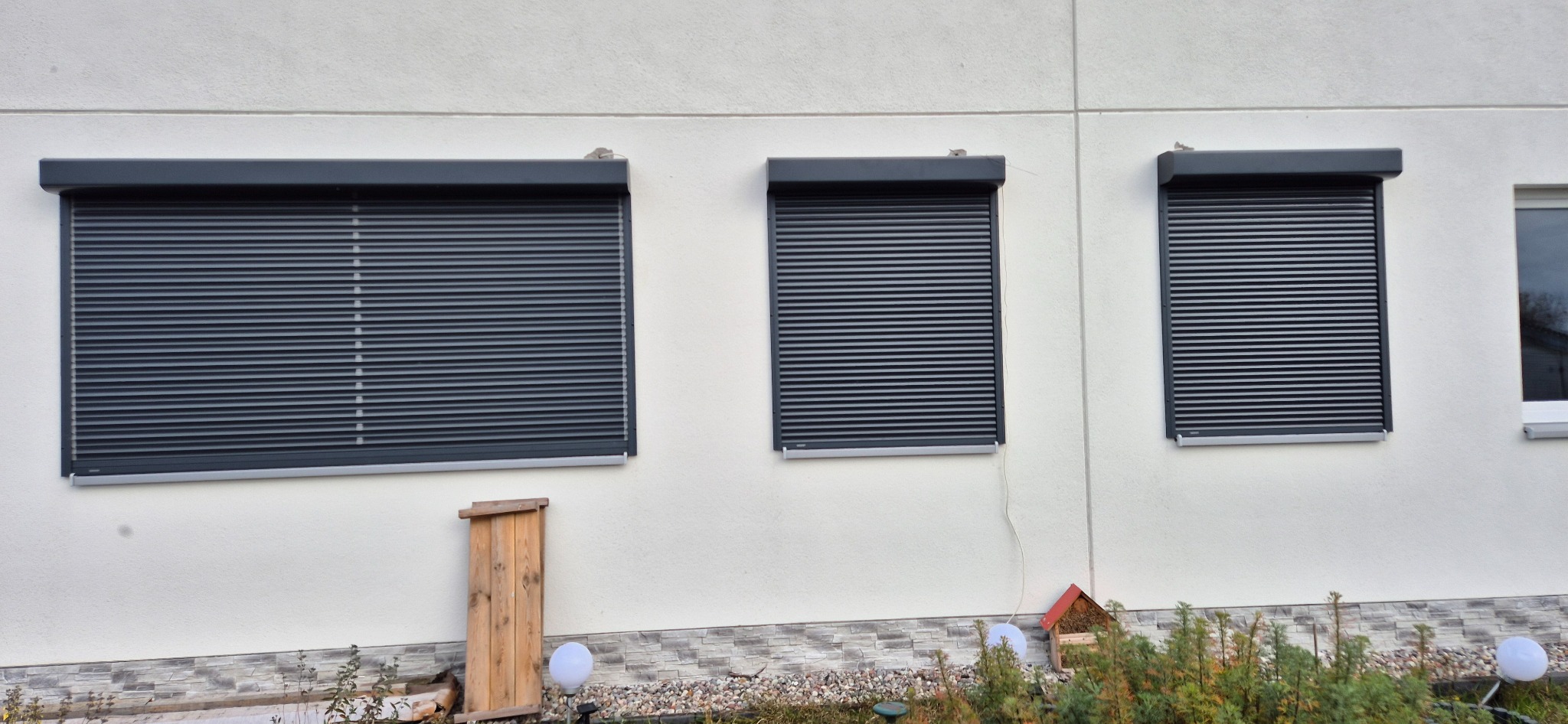 Elewacja domu z trzema oknami zabezpieczonymi szarymi roletami zewnętrznymi, widoczne elementy dekoracyjne ogrodu.