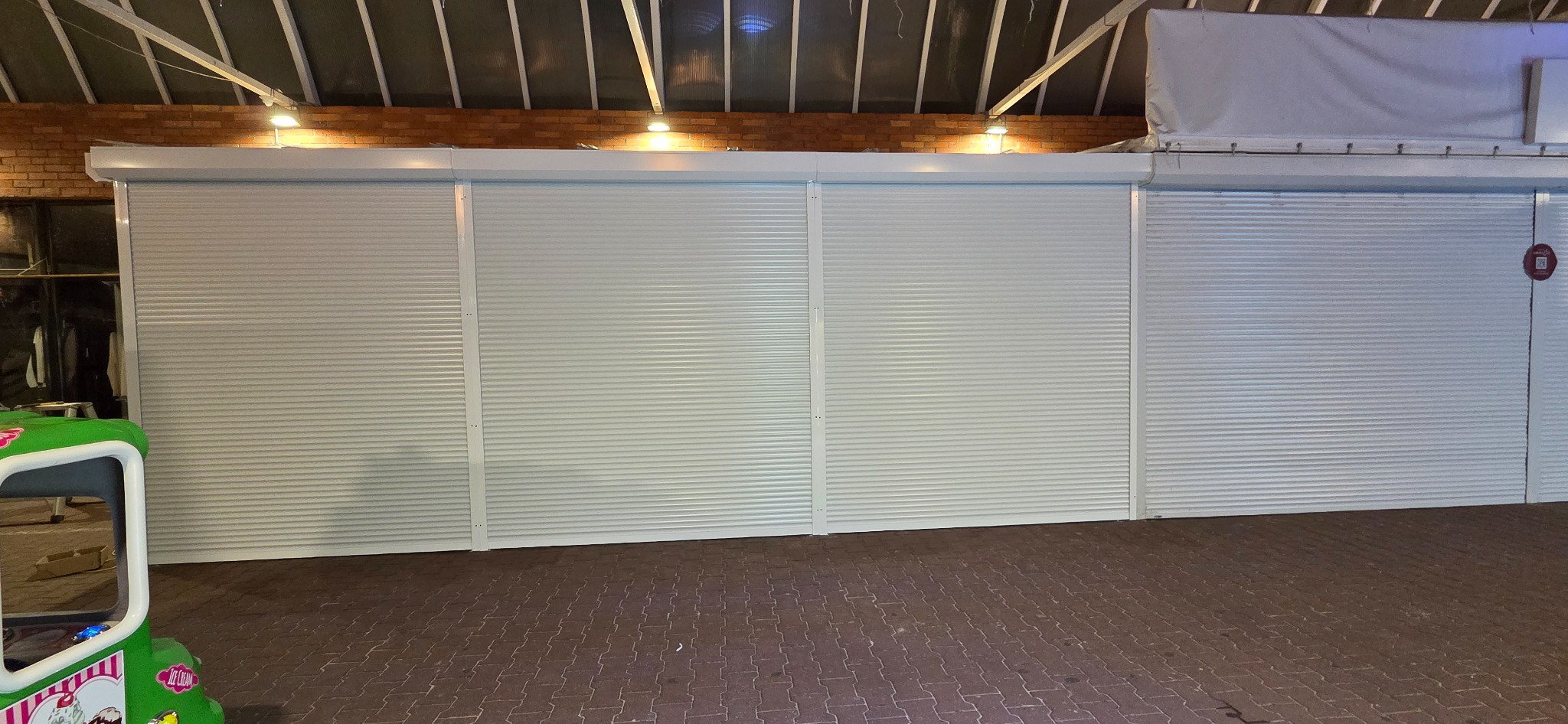 Zewnętrzne, białe rolety zabezpieczające witrynę sklepową, widoczne z zewnątrz na tle ceglanej ściany i kostki brukowej.