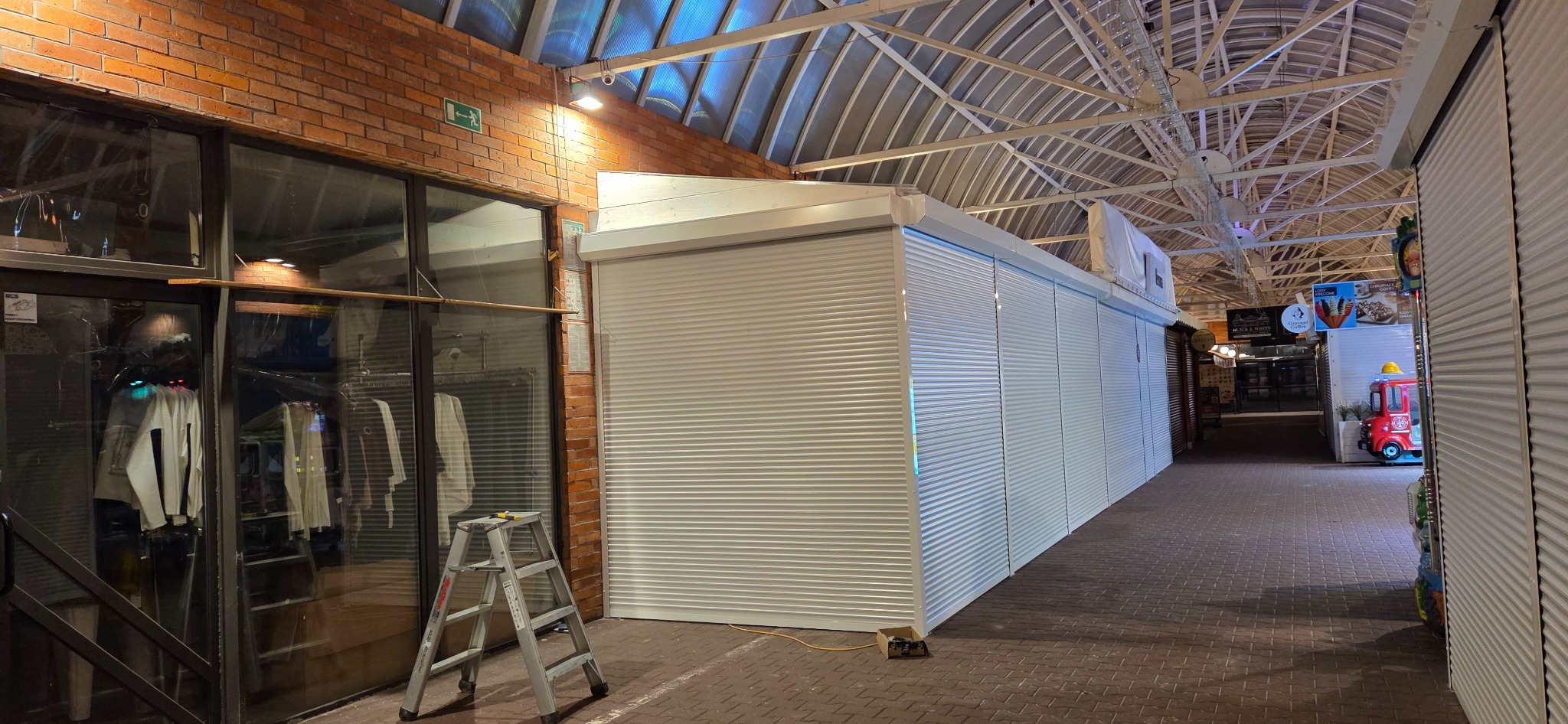 Zasunięte, białe rolety na stoiskach w hali targowej, widok z perspektywy alejki. Aluminiowa drabina oparta o ścianę z cegły. Oświetlenie sufitowe.