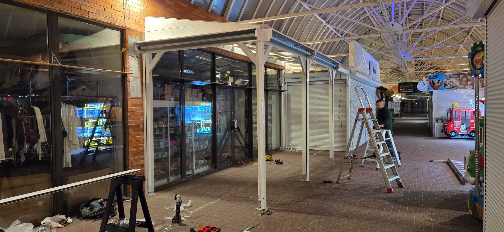 Montaż białej konstrukcji z elementami zwiniętej osłony na zewnątrz lokalu w pasażu handlowym. Widoczne narzędzia i drabina.