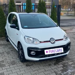Biały Volkswagen Up! GTI zaparkowany na kostce brukowej przed nowoczesnym budynkiem z szarymi panelami i rzędem zielonych tuj, widoczne czarne felgi i czerwony akcent na grillu.