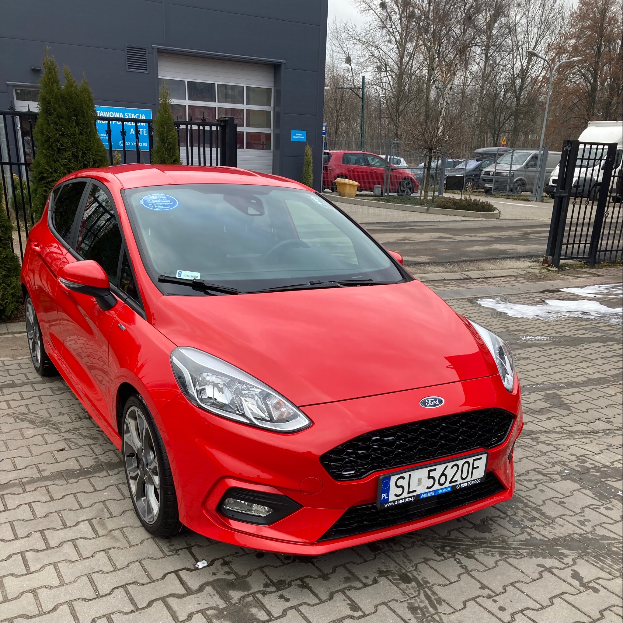 Czerwony Ford Fiesta po myjni, zaparkowany przed budynkiem stacji mycia pojazdów z widoczną tablicą rejestracyjną SL 5620F i naklejką informacyjną na szybie.