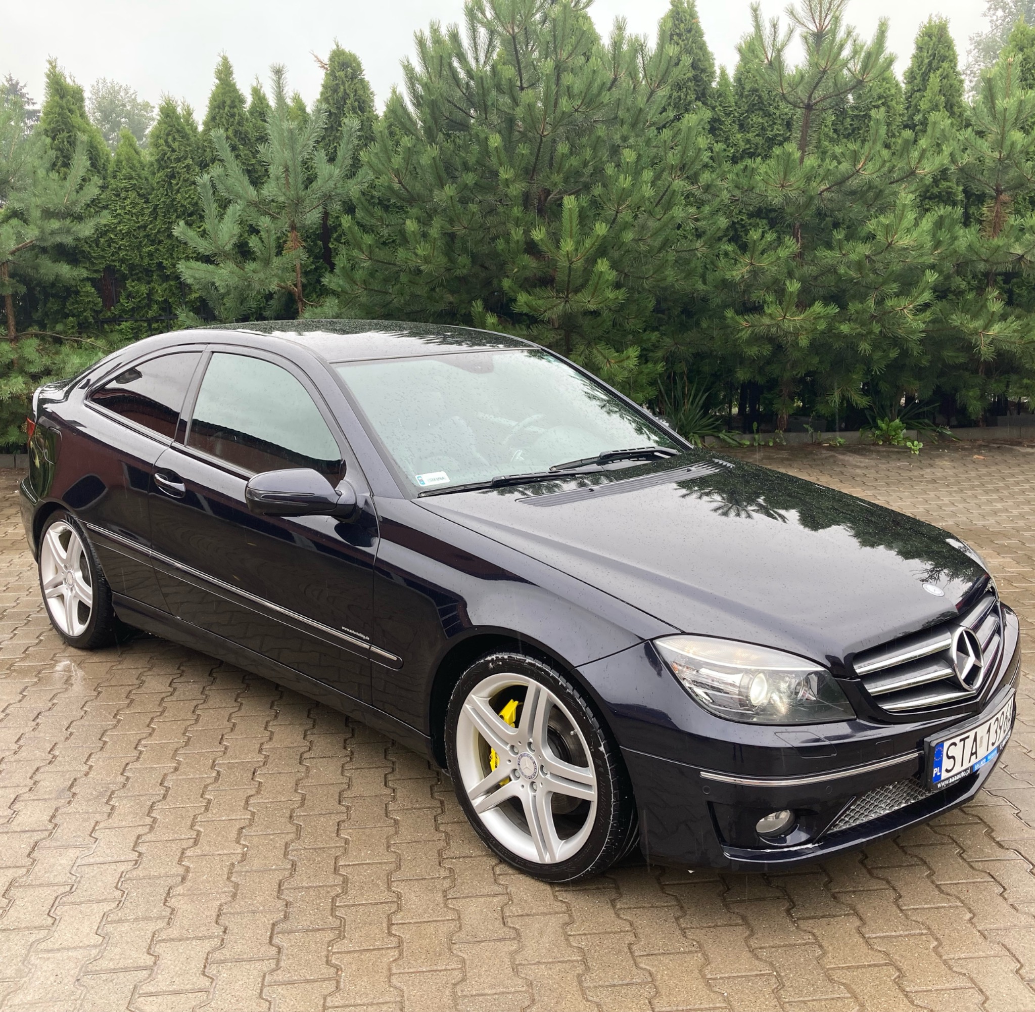 Ciemny samochód Mercedes CLC zaparkowany na mokrej kostce brukowej, z widocznymi żółtymi zaciskami hamulcowymi i zielonym tłem z drzew iglastych.