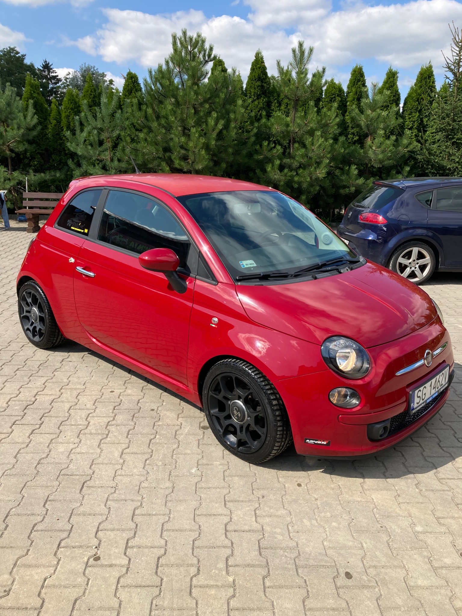Czerwony Fiat 500 z czarnymi felgami stoi na kostce brukowej, na tle zielonych drzew iglastych i zaparkowanego niebieskiego samochodu.