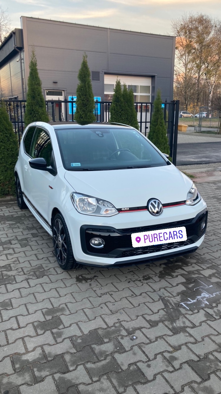 Biały Volkswagen Up! GTI zaparkowany na kostce brukowej przed nowoczesnym budynkiem z szarymi panelami i rzędem zielonych tuj, widoczne czarne felgi i czerwony akcent na grillu.