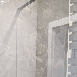 Mp-Perfekt - Ściana wyłożona szarymi, heksagonalnymi płytkami z delikatnym wzorem, obok widoczny fragment grzejnika łazienkowego za szklanym panelem.