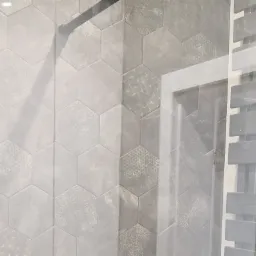 Ściana wyłożona szarymi, heksagonalnymi płytkami z delikatnym wzorem, obok widoczny fragment grzejnika łazienkowego za szklanym panelem.