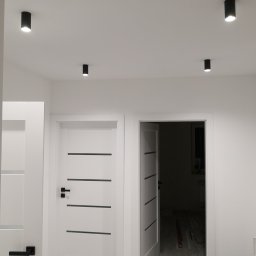 Mp-Perfekt - Nowoczesne wnętrze w bieli z minimalistycznymi czarnymi lampami sufitowymi, widoczne dwie białe drzwi z czarnymi klamkami i poziomymi wstawkami, jedna otwarta ukazująca fragment wnętrza.