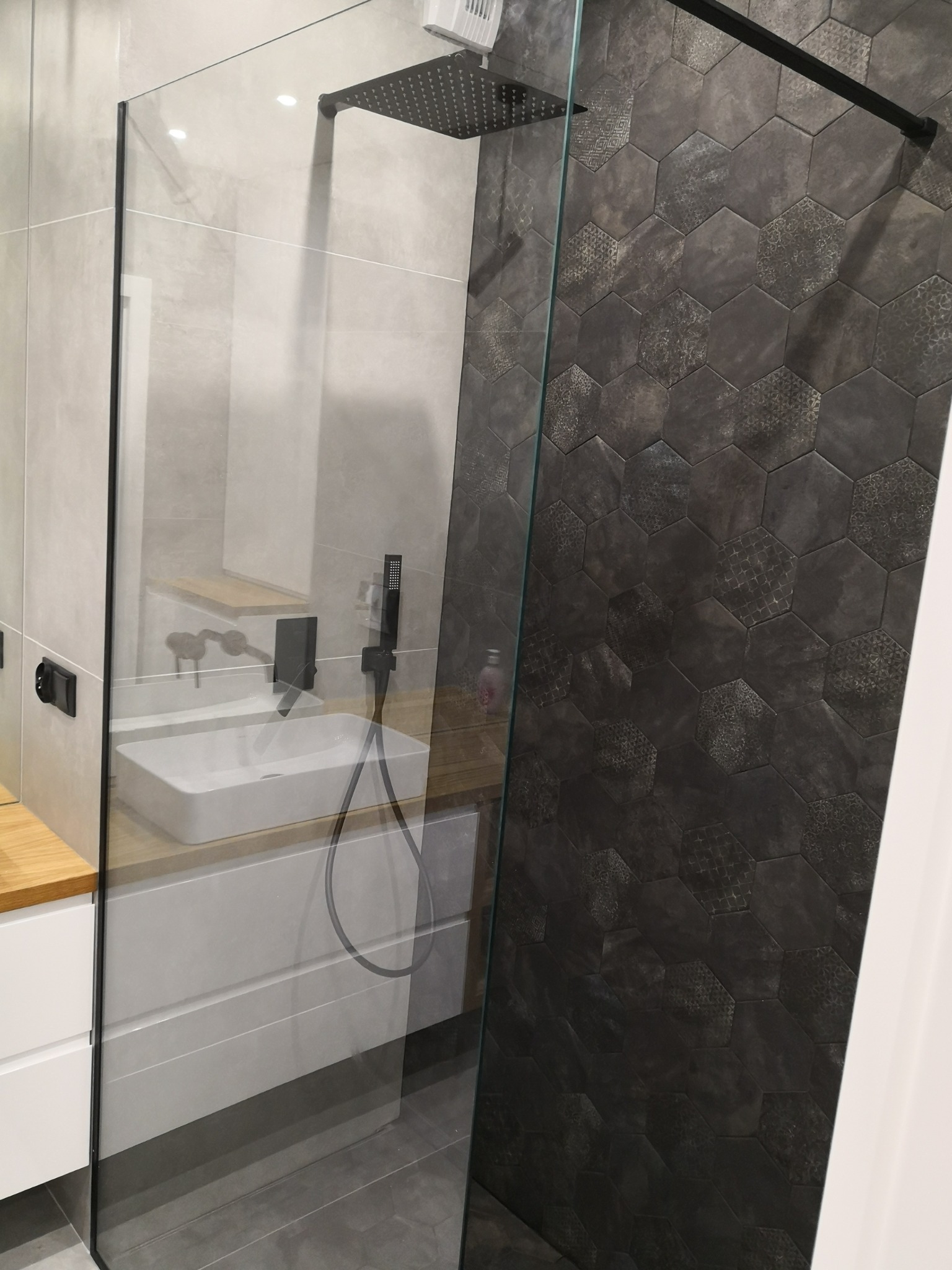 Nowoczesna łazienka z kabiną prysznicową typu walk-in, heksagonalnymi płytkami w odcieniach szarości, białą umywalką nablatową na drewnianym blacie i czarną armaturą.
