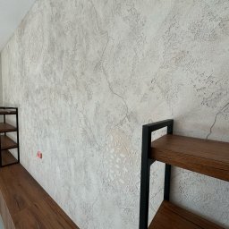 WAŻKA Usługi Budowlane - Ściana wykończona dekoracyjną gładzią imitującą spękania i zdobiona ornamentami, z fragmentem drewnianego regału z czarnymi metalowymi elementami po bokach.