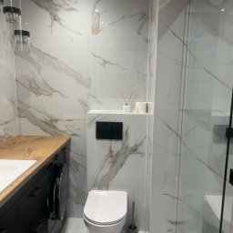 WAŻKA Usługi Budowlane - Nowoczesna łazienka z białymi płytkami imitującymi marmur, czarną szafką z drewnianym blatem, wiszącą toaletą i szklaną kabiną prysznicową, oświetlona wiszącymi lampami.