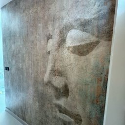 WAŻKA Usługi Budowlane - Ściana z fototapetą przedstawiającą profil twarzy o fakturze starego tynku, widoczny fragment białej listwy przypodłogowej i włącznik światła.