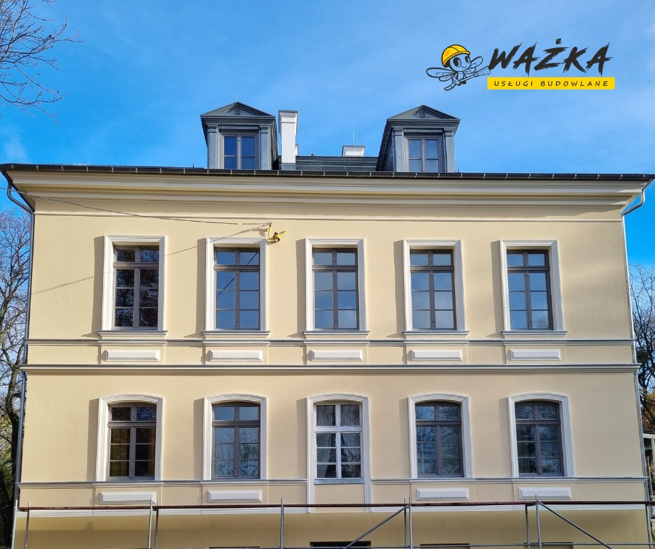 Odnowiona elewacja dwupiętrowego budynku z szarą stolarką okienną i poddaszem, widoczne rusztowanie na pierwszym planie oraz logo firmy budowlanej w prawym górnym rogu.