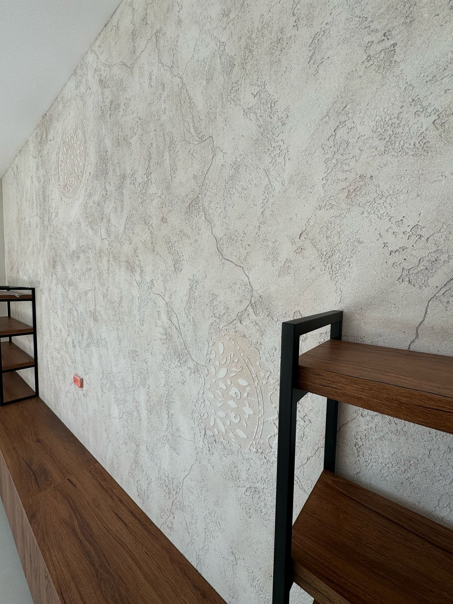 Ściana wykończona dekoracyjną gładzią imitującą spękania i zdobiona ornamentami, z fragmentem drewnianego regału z czarnymi metalowymi elementami po bokach.