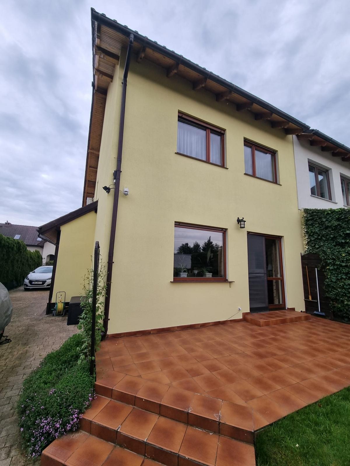 Żółta elewacja dwukondygnacyjnego domu z brązowymi ramami okiennymi, widoczny taras z brązowej terakoty i fragment podjazdu z samochodem w tle.