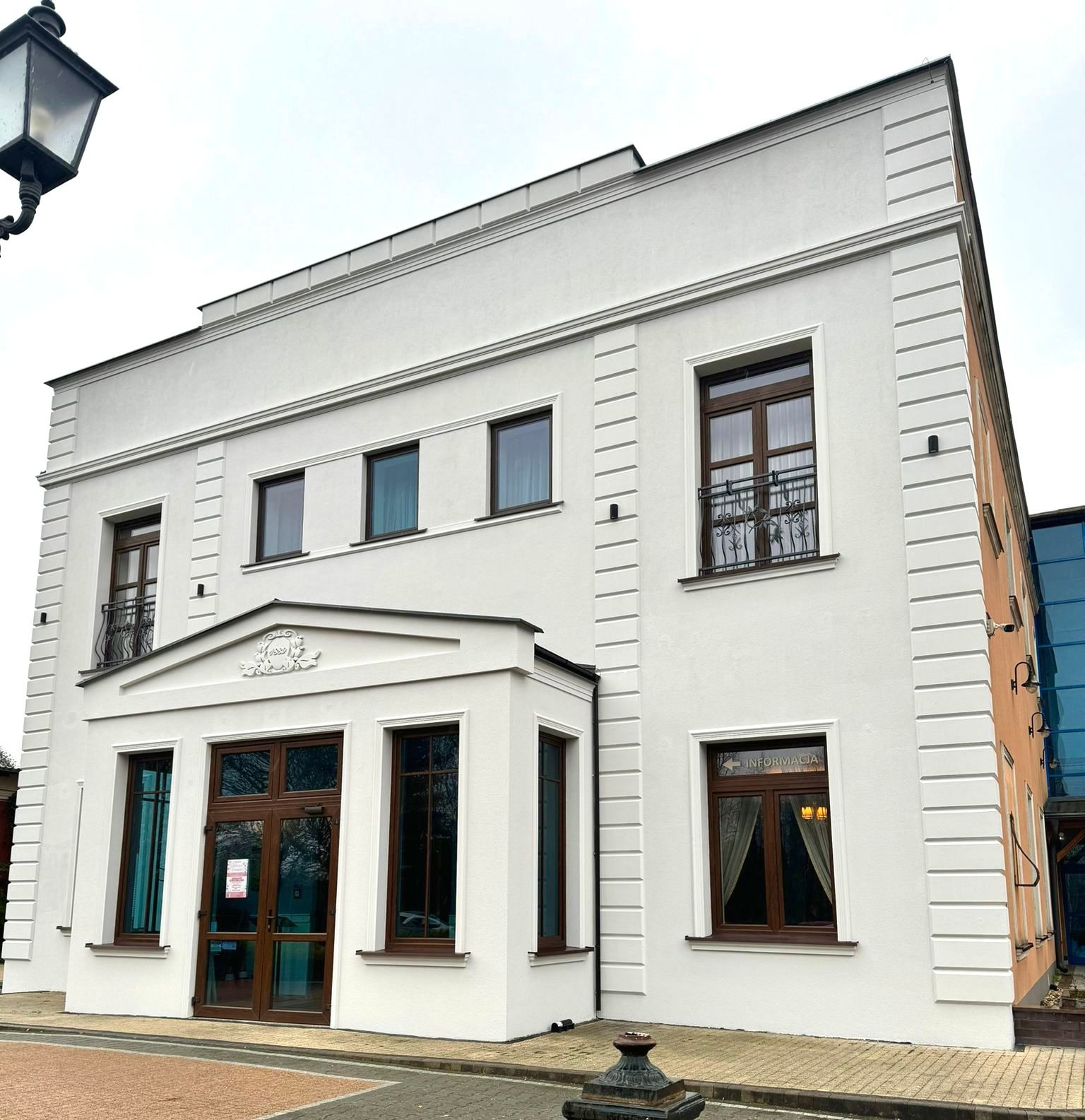 Biała elewacja odnowionego budynku z brązowymi oknami, ozdobnym balkonem i napisem 'Informacja' w oknie, widziana z poziomu chodnika. W tle błękitny fragment nowszej dobudówki.